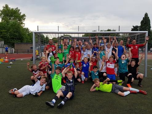 Gruppenshot von Kindern in Sportkleidung vor einem Fußballtor, jubelnd mit erhobenen Händen auf einem Kunstrasenplatz.