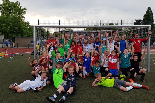 Gruppenshot von Kindern in Sportkleidung vor einem Fußballtor, jubelnd mit erhobenen Händen auf einem Kunstrasenplatz.