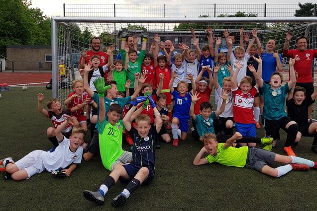 Gruppenshot von Kindern in Sportkleidung vor einem Fu&szlig;balltor, jubelnd mit erhobenen H&auml;nden auf einem Kunstrasenplatz.
