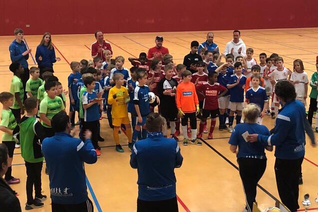 Gruppierung von Kindern in Fu&szlig;balltrikots nach einem Turnier, applaudiert von Trainern in einer Sporthalle.
