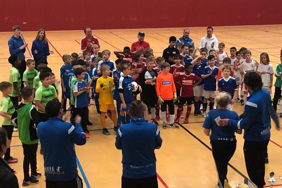 Gruppierung von Kindern in Fußballtrikots nach einem Turnier, applaudiert von Trainern in einer Sporthalle.