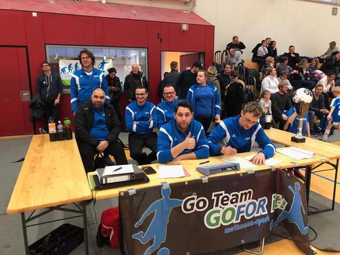 Sechs Personen in Sportjacken sitzen an einem Tisch mit Pokal und Getränken in einer Sporthalle voller Zuschauer.