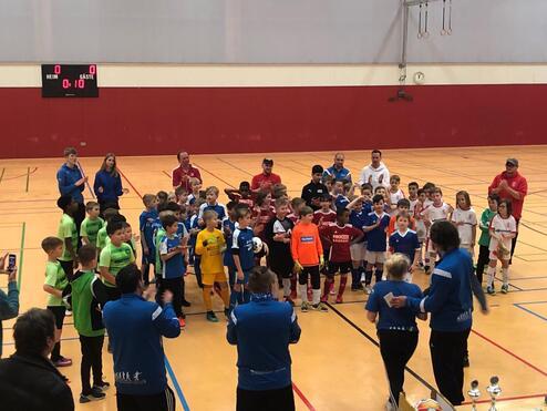 Gruppierung von Kindern in Fußballtrikots nach einem Turnier, applaudiert von Trainern in einer Sporthalle.