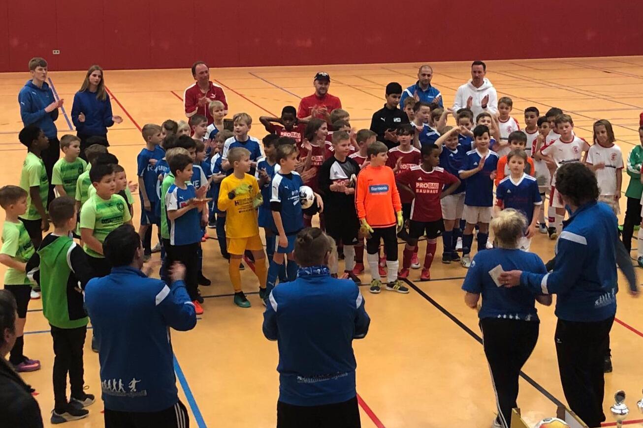 Gruppierung von Kindern in Fu&szlig;balltrikots nach einem Turnier, applaudiert von Trainern in einer Sporthalle.