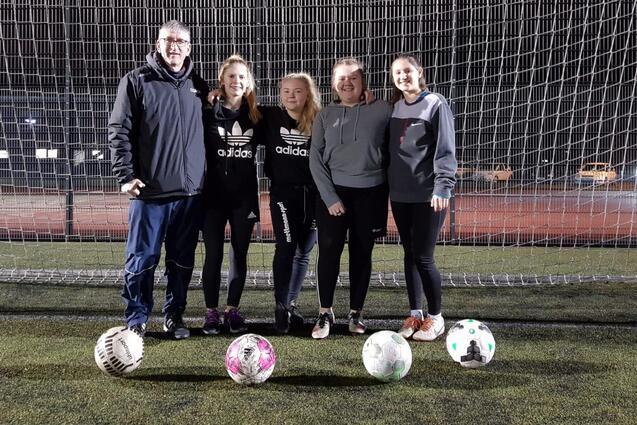 F&uuml;nf Personen, darunter ein Trainer, stehen vor einem Fu&szlig;balltor mit mehreren Fu&szlig;ballen auf einem Kunstrasenplatz.