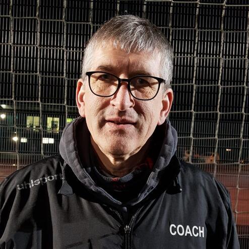 Mann mit Brille und grauen Haaren trägt eine schwarze Jacke mit dem Aufdruck „COACH“ vor einem Fußballtor bei Nacht.
