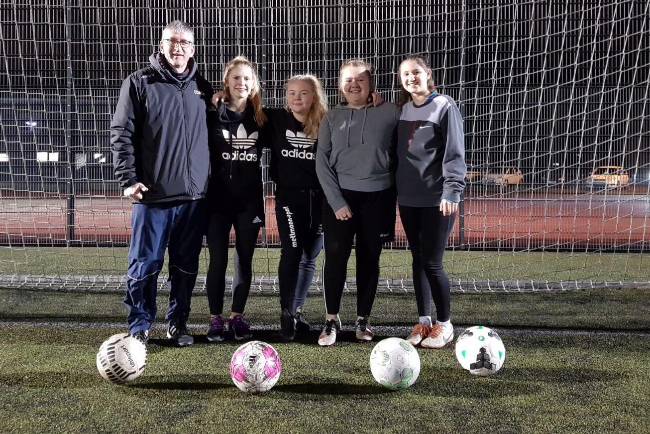 F&uuml;nf Personen, darunter ein Trainer, stehen vor einem Fu&szlig;balltor mit mehreren Fu&szlig;ballen auf einem Kunstrasenplatz.