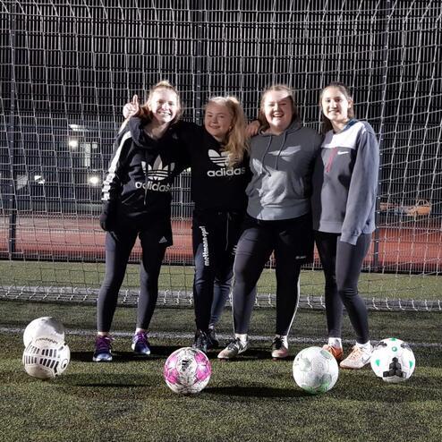 Vier junge Frauen stehen vor einem Fußballtor, während drei Bälle auf dem Kunstrasen liegen.