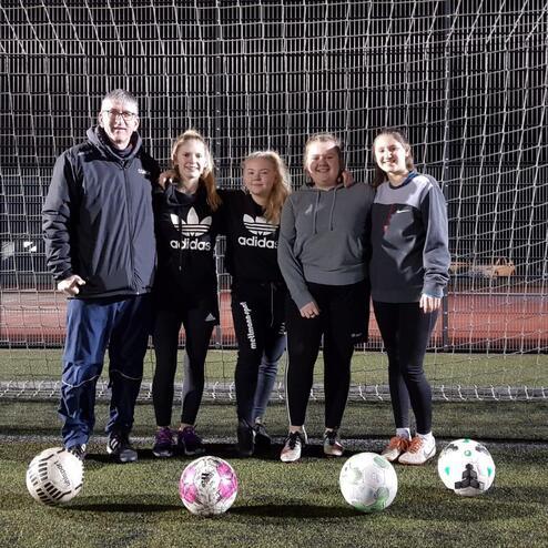 Fünf Personen, darunter ein Trainer, stehen vor einem Fußballtor mit mehreren Fußballen auf einem Kunstrasenplatz.