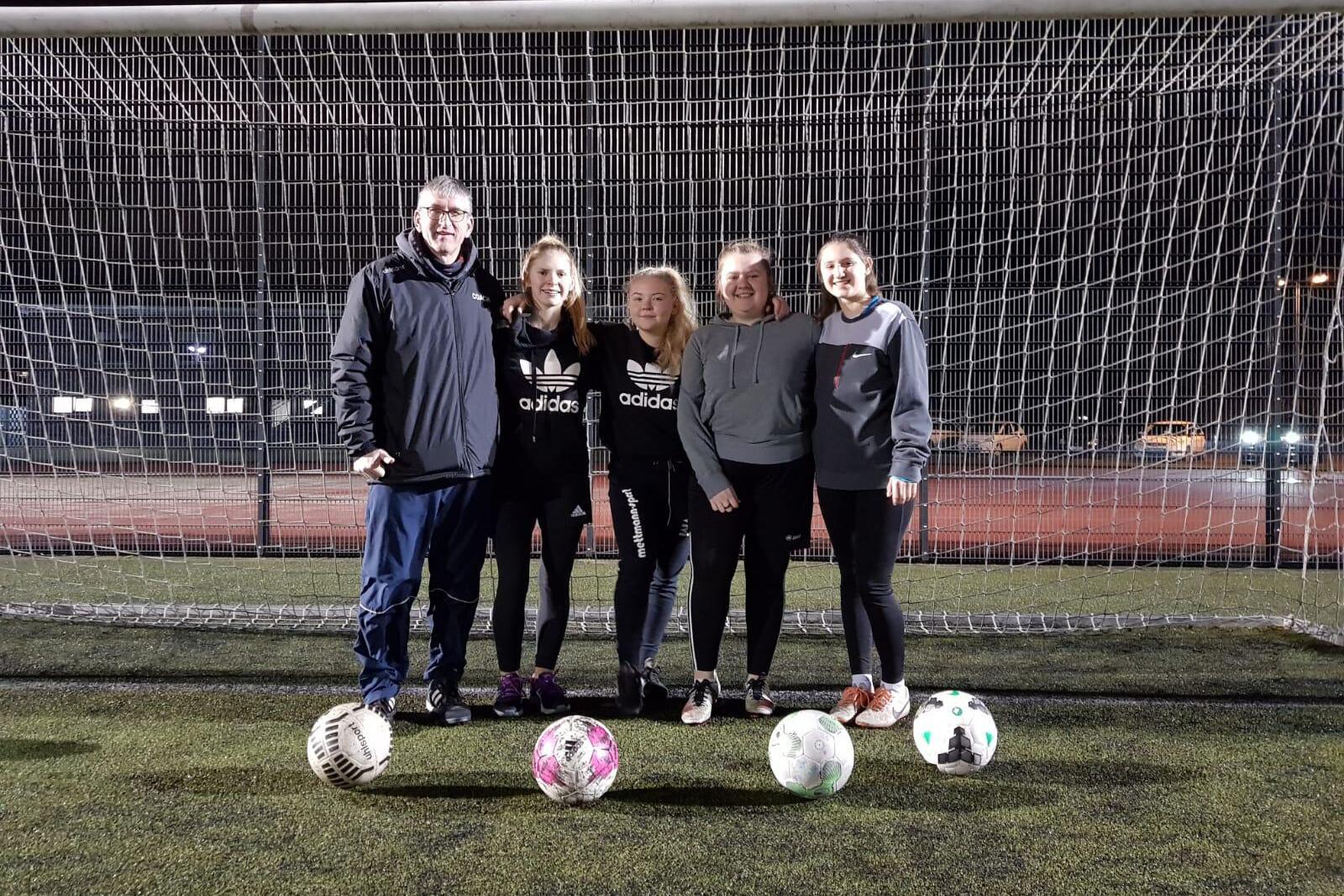 Fünf Personen, darunter ein Trainer, stehen vor einem Fußballtor mit mehreren Fußballen auf einem Kunstrasenplatz.