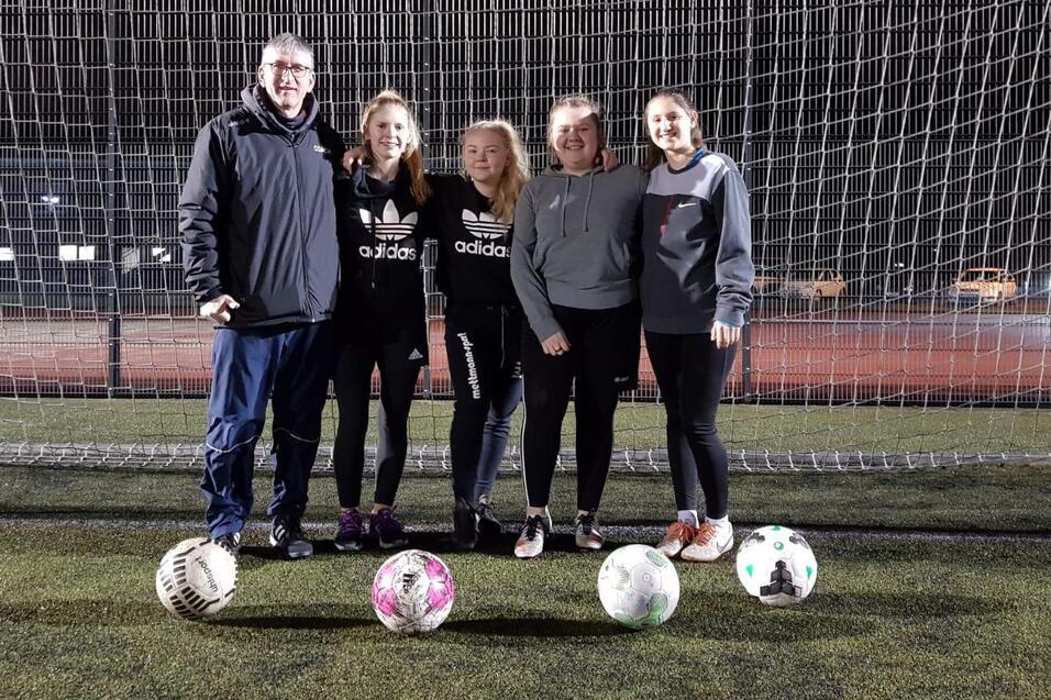 Fünf Personen, darunter ein Trainer, stehen vor einem Fußballtor mit mehreren Fußballen auf einem Kunstrasenplatz.
