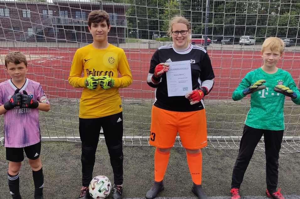 Vier Fußballtorhüter in Ausrüstung stehen vor einem Tor, eine Person hält ein Zertifikat in der Hand.