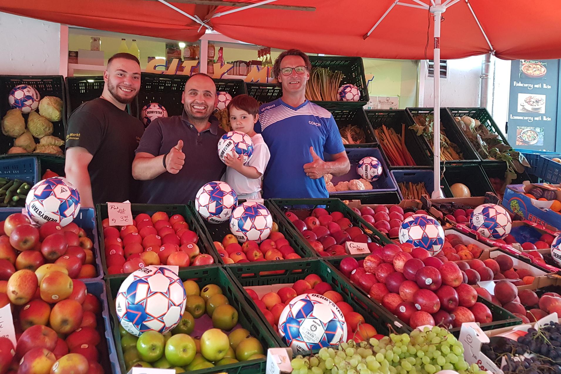 Vier M&auml;nner und ein Junge l&auml;cheln in einem Marktstand zwischen Obstkisten mit Fu&szlig;ball-Motiven in der Auslage.
