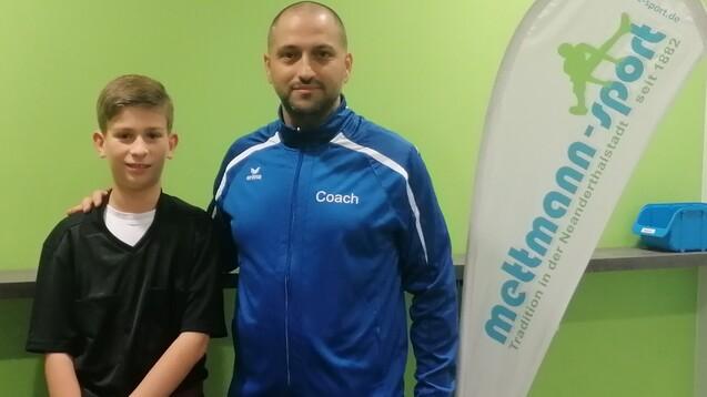 Ein Junge in Sportkleidung steht neben einem Trainer in blauer Trainerjacke vor einem grünen Hintergrund.