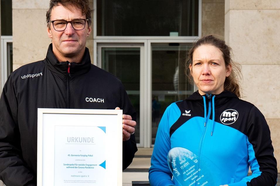 Zwei Personen halten Urkunden und Auszeichnungen im Freien vor einem Gebäude. Die Person links trägt eine schwarze Jacke, die rechte eine blaue.