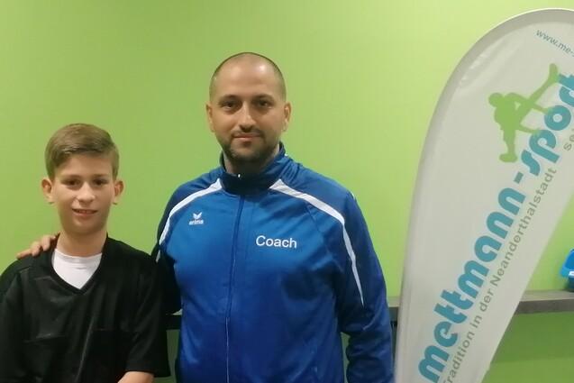 Ein Junge in Sportkleidung steht neben einem Trainer in blauer Trainerjacke vor einem gr&uuml;nen Hintergrund.