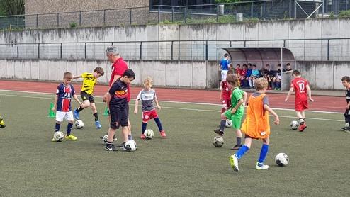 Kinder in Fußballtrikots trainieren auf einem Kunstrasenplatz, während ein Trainer Anweisungen gibt.