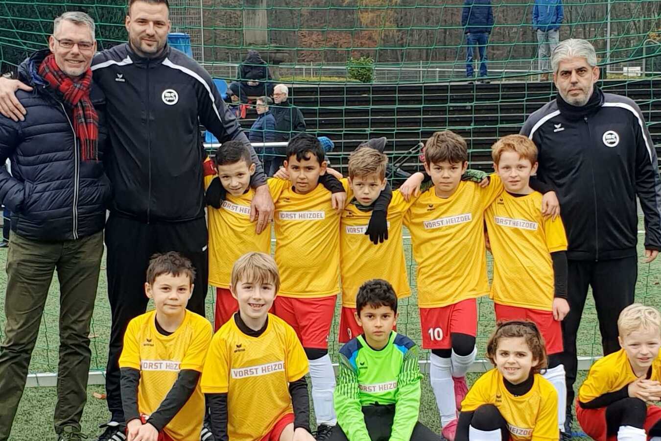 Gruppenfoto einer Jugendfu&szlig;ballmannschaft mit zwei Trainern, alle in gelben Trikots, vor einem Fu&szlig;balltor.