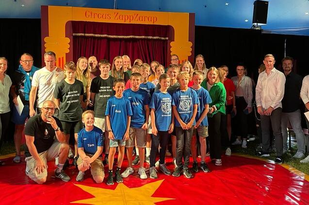 Gruppensfoto von Kindern und Erwachsenen im Zirkuszelt vor einer B&uuml;hne mit dem Schriftzug &bdquo;Circus ZappZarap&ldquo;.