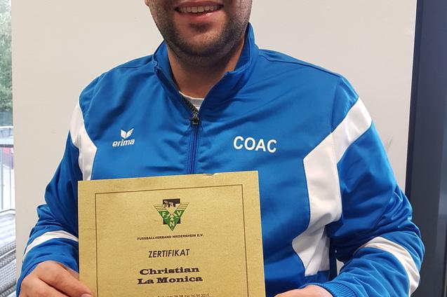 Mann in blauer Sportjacke hält stolz ein Zertifikat für Kindertraining in der Fußballeinrichtung.