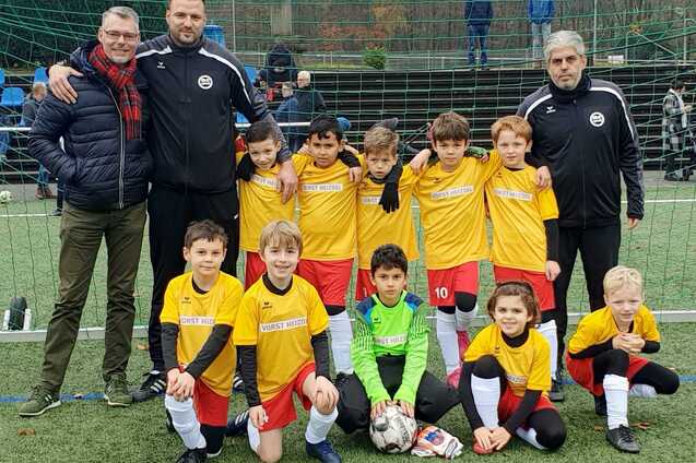 Gruppenfoto einer Jugendfußballmannschaft mit zwei Trainern, alle in gelben Trikots, vor einem Fußballtor.
