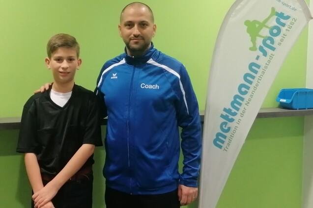 Ein Junge in Sportkleidung steht neben einem Trainer in blauer Trainerjacke vor einem grünen Hintergrund.