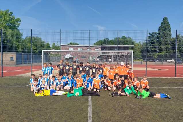 Gruppenfoto von Kindern in blauen und orangefarbenen Fußballtrikots auf einem Sportplatz vor einem Fußballtor.