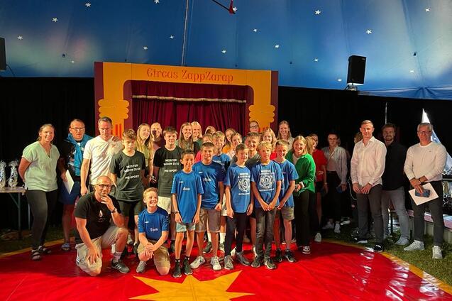 Gruppensfoto von Kindern und Erwachsenen im Zirkuszelt vor einer Bühne mit dem Schriftzug „Circus ZappZarap“.