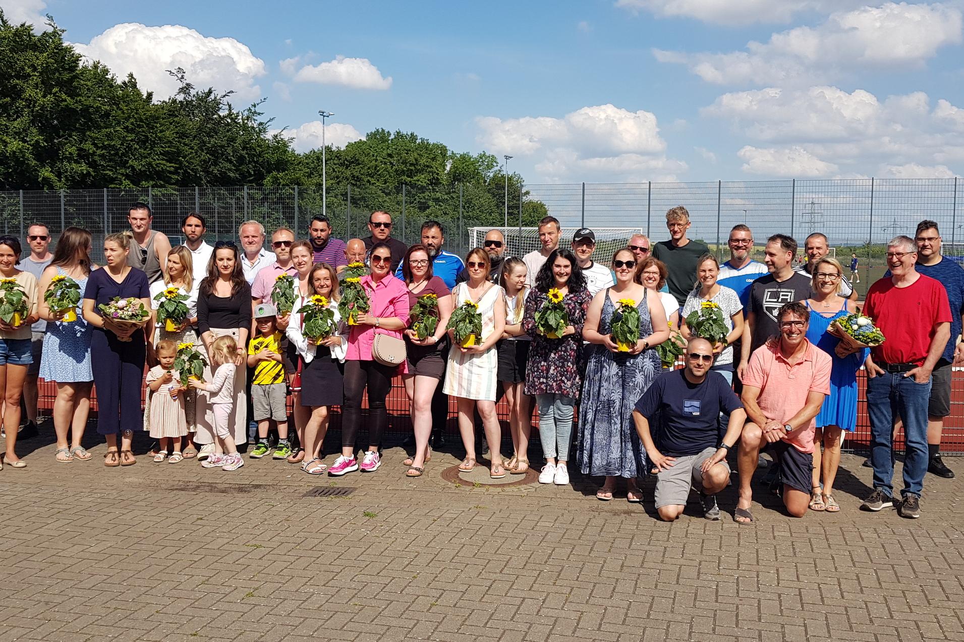 Gruppe von Personen mit Blumenstr&auml;u&szlig;en auf einem Sportplatz, darunter Kinder, bei sonnigem Wetter.