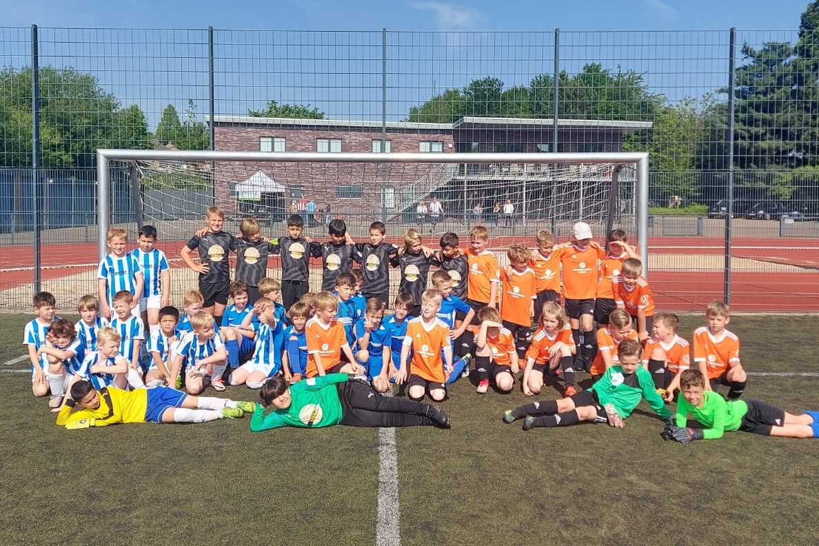Gruppenfoto von Kindern in blauen und orangefarbenen Fu&szlig;balltrikots auf einem Sportplatz vor einem Fu&szlig;balltor.