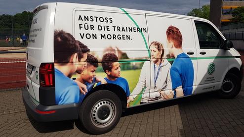 Fahrzeug mit Werbebotschaft für Trainerinnen, umgeben von Jugendlichen in Sportkleidung auf dem Spielfeld.