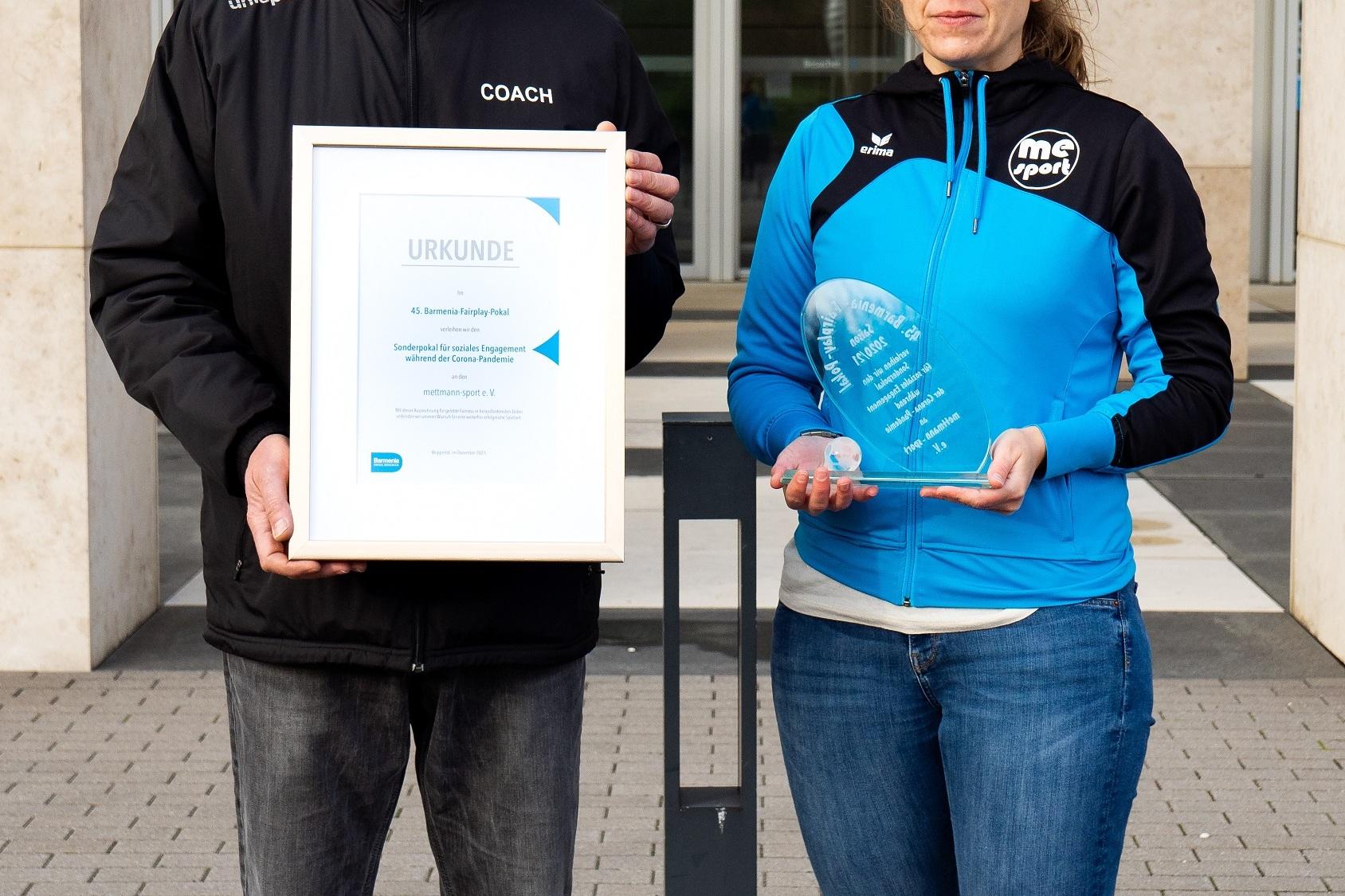 Zwei Personen halten Urkunden und Auszeichnungen im Freien vor einem Gebäude. Die Person links trägt eine schwarze Jacke, die rechte eine blaue.