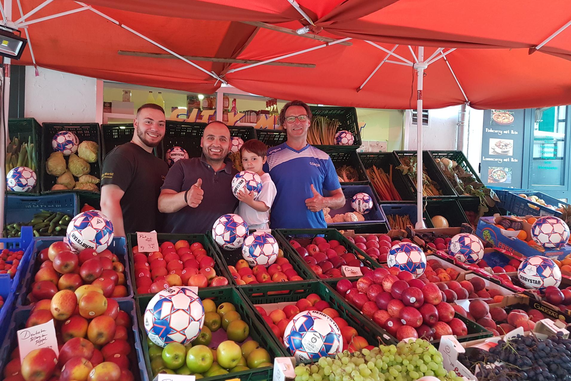 Vier Männer und ein Junge lächeln in einem Marktstand zwischen Obstkisten mit Fußball-Motiven in der Auslage.