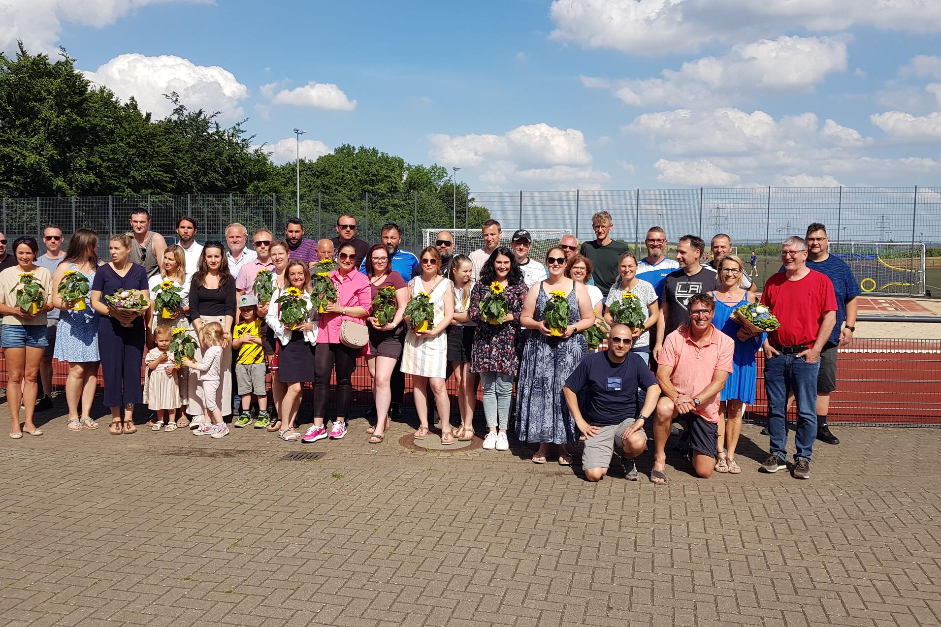 Gruppe von Personen mit Blumensträußen auf einem Sportplatz, darunter Kinder, bei sonnigem Wetter.