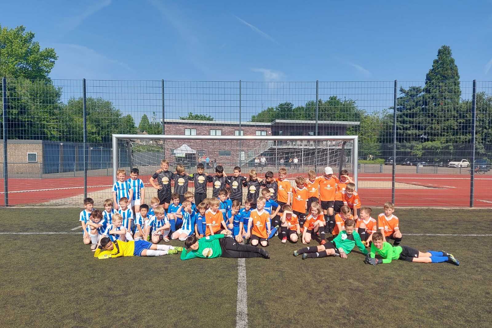 Gruppenfoto von Kindern in blauen und orangefarbenen Fußballtrikots auf einem Sportplatz vor einem Fußballtor.