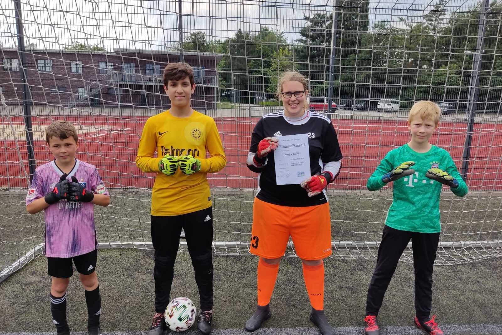 Vier Fußballtorhüter in Ausrüstung stehen vor einem Tor, eine Person hält ein Zertifikat in der Hand.