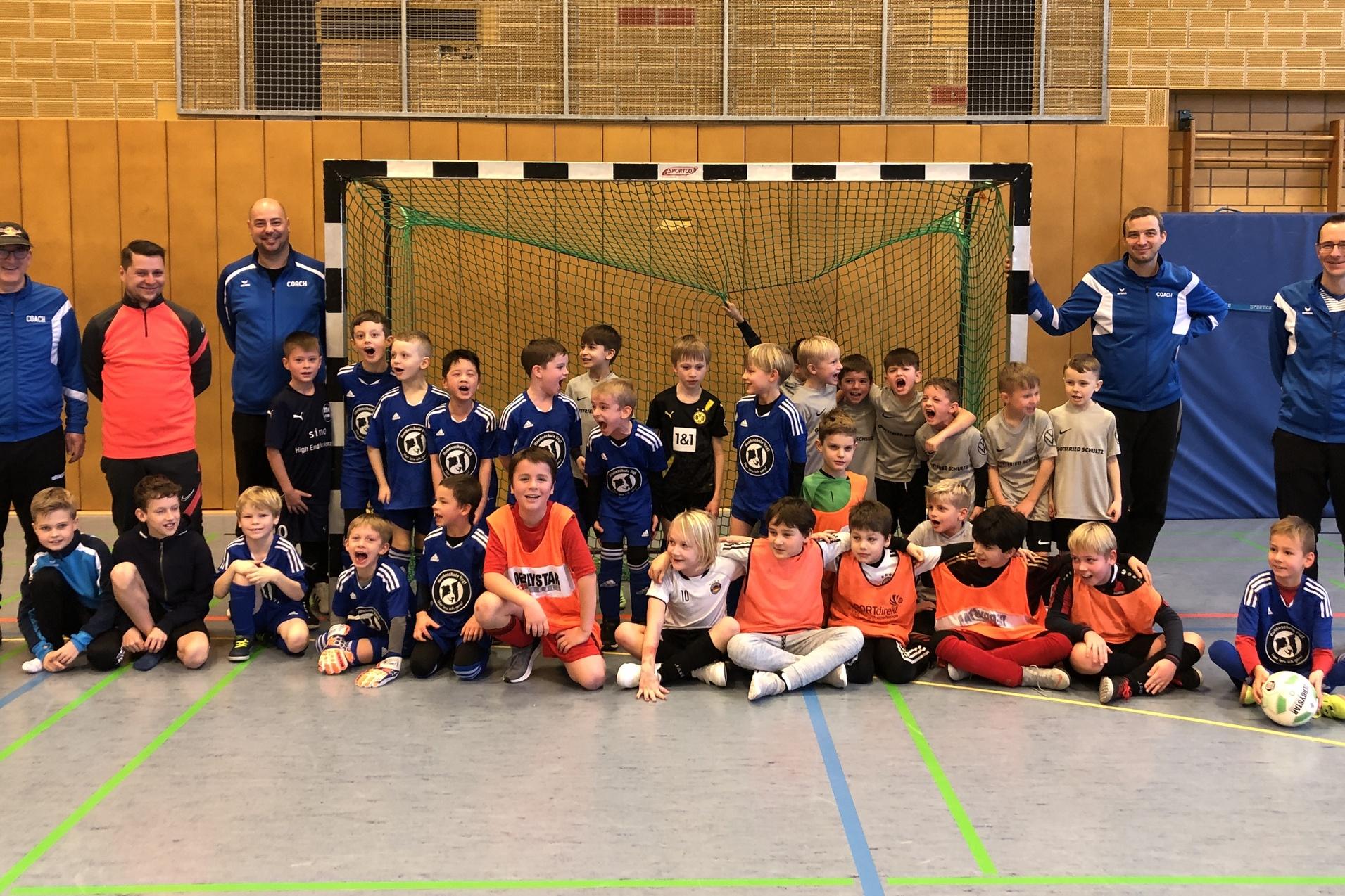 Gruppenfoto mit Kindern und Trainern in Sportkleidung vor einem Fu&szlig;balltor in einer Sporthalle.