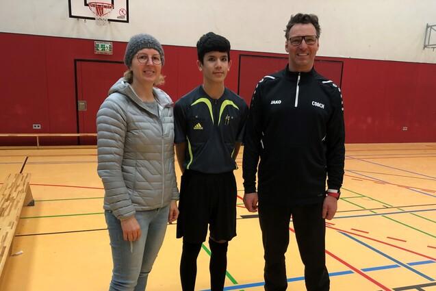 Drei Personen stehen in einer Sporthalle, eine Frau und zwei Männer in Sportkleidung, davor eine Bank und ein Basketballkorb.