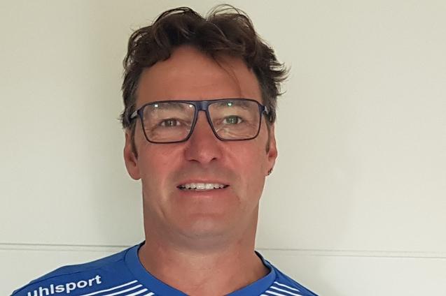 Mann in blauer Sportkleidung h&auml;lt ein Zertifikat in der Hand, l&auml;chelt und steht vor einer hellen Wand.