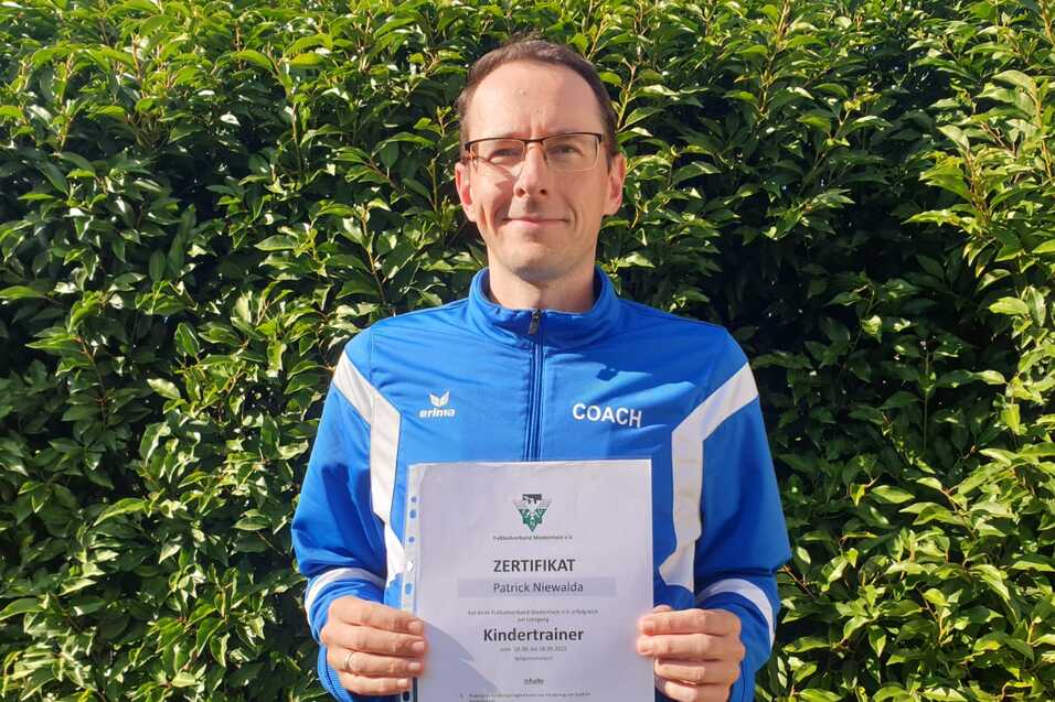 Mann hält ein Zertifikat für die Trainerausbildung zum Kindertrainer vor einer grünen Hecke.