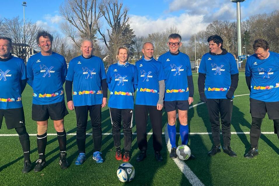 Gruppenfoto von sieben Fußballspielern in blauen Trikots auf einem grünen Platz mit Bäumen und blauem Himmel im Hintergrund.