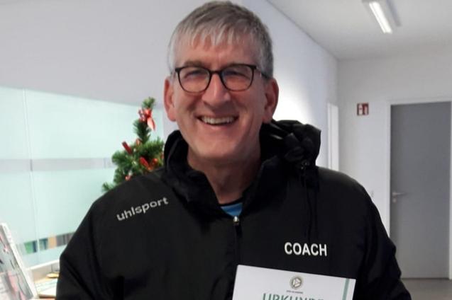 Mann in schwarzer Jacke h&auml;lt stolz eine Trainerurkunde in der Hand, mit einem L&auml;cheln im modernen Raum.