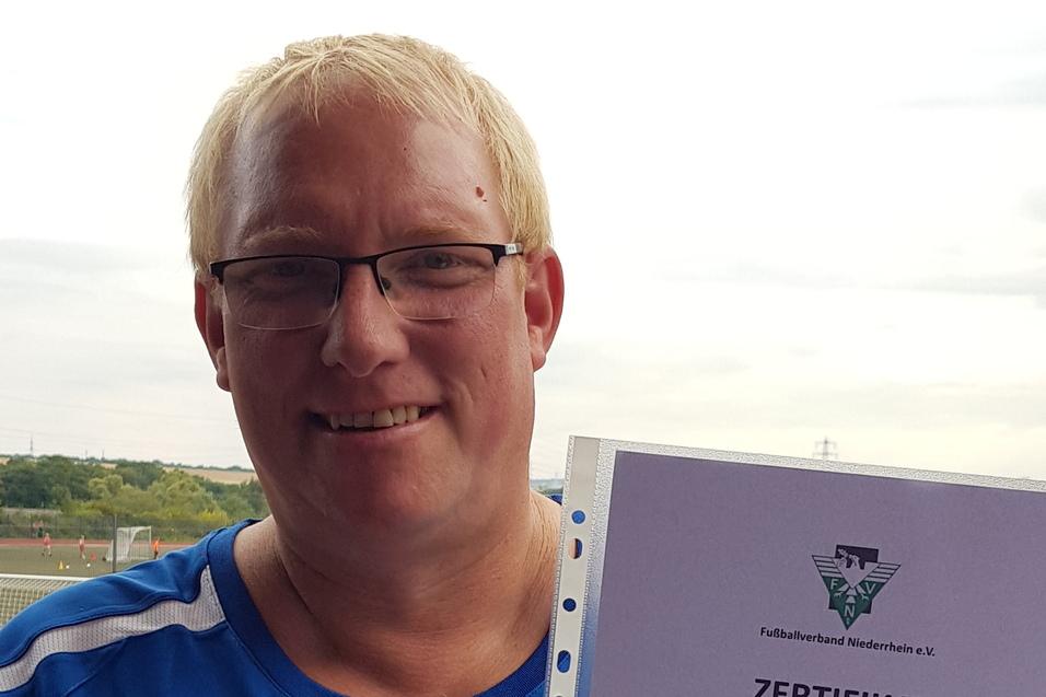 Mann hält ein Zertifikat in der Hand, das ihn als Kindertrainer ausweist, vor einem Sportplatz.