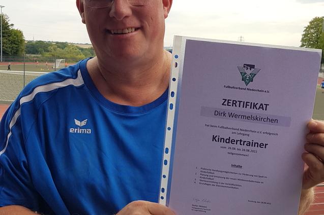Mann hält ein Zertifikat in der Hand, das ihn als Kindertrainer ausweist, vor einem Sportplatz.