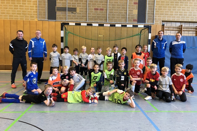 Gruppenfoto von Kindern in Fußballtrikots und Trainern vor einem Tor in einer Sporthalle.