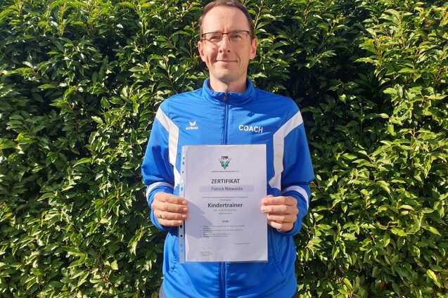 Mann hält ein Zertifikat für die Trainerausbildung zum Kindertrainer vor einer grünen Hecke.