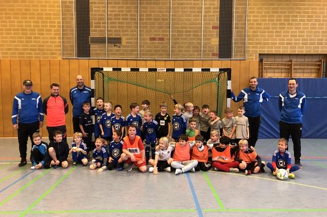 Gruppenfoto mit Kindern und Trainern in Sportkleidung vor einem Fußballtor in einer Sporthalle.