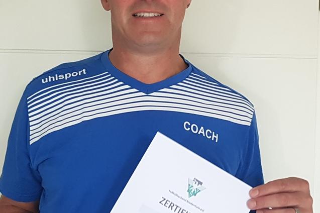 Mann in blauer Sportkleidung hält ein Zertifikat in der Hand, lächelt und steht vor einer hellen Wand.