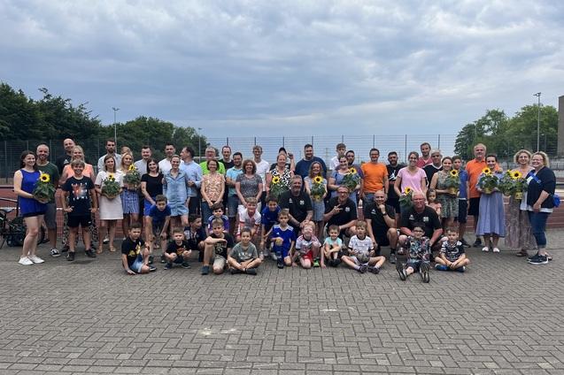 Gruppe von Personen unterschiedlichen Alters mit Sonnenblumen in der Hand, versammelt auf einem Platz nahe einem Tennisplatz.