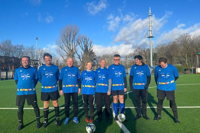 Gruppenfoto von sieben Fußballspielern in blauen Trikots auf einem grünen Platz mit Bäumen und blauem Himmel im Hintergrund.